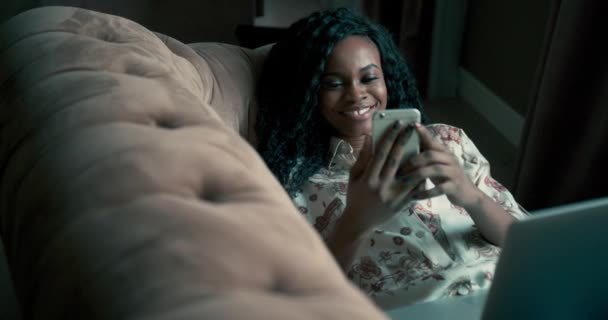 Hermosa mujer afroamericana con un peinado afro muy rizado mensajes de texto en su móvil, mientras que ella acostada en un sofá. Mujer joven ver algo en su teléfono y divertirse. Increíble chica de exótico — Vídeo de stock