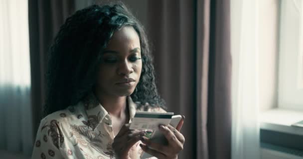 Όμορφη Αφροαμερικανή γυναίκα με ένα κατσαρό afro χτένισμα αγοράζοντας κάτι σε απευθείας σύνδεση χρησιμοποιώντας το κινητό και την πιστωτική της κάρτα. Αγορές σε απευθείας σύνδεση από χαρούμενα όμορφη μελαχρινή. Βίντεο 4K — Αρχείο Βίντεο