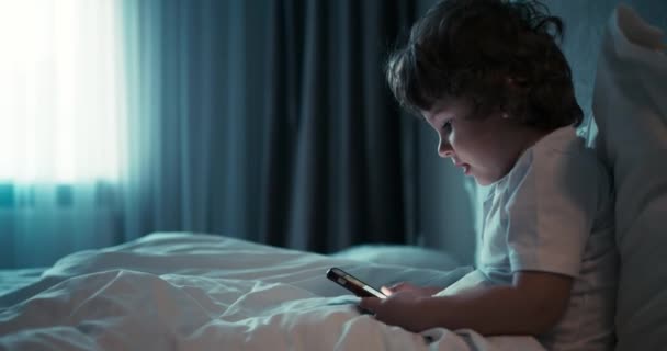 Un bambino è seduto sul letto e utilizza uno smartphone. Il bambino guarda i cartoni animati su uno smartphone prima di coricarsi. — Video Stock