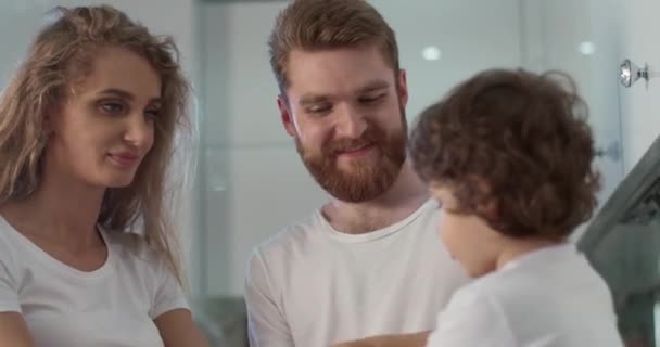 Bom dia em casa. Jovens pais brincando com seu adorável filho bebê no interior da cozinha. Eles colocaram uma mão na mão e levantaram. Cozinha acolhedora branca e ambiente familiar calmo. Vídeo 4K — Vídeo de Stock