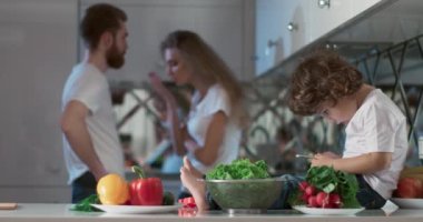 Oğulları yanlarında oturmuş telefonda oyun oynarken mutfakta tartışan genç bir çiftin portresi. Ayrıca ön planda farklı sebzeler ve meyveler vardır. 4K