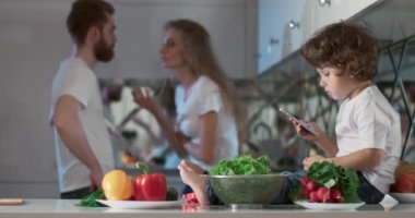 Oğulları yanlarında oturmuş telefonda oyun oynarken mutfakta tartışan genç bir çiftin portresi. Ayrıca ön planda farklı sebzeler ve meyveler vardır. 4K