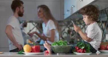 Oğulları yanlarında oturmuş telefonda oyun oynarken mutfakta tartışan genç bir çiftin portresi. Ayrıca ön planda farklı sebzeler ve meyveler vardır. 4K