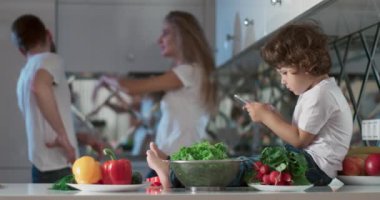 Oğulları yanlarında oturmuş telefonda oyun oynarken mutfakta tartışan genç bir çiftin portresi. Ayrıca ön planda farklı sebzeler ve meyveler vardır. 4K