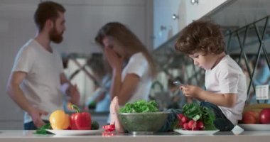 Oğulları yanlarında oturmuş telefonda oyun oynarken mutfakta tartışan genç bir çiftin portresi. Ayrıca ön planda farklı sebzeler ve meyveler vardır. 4K