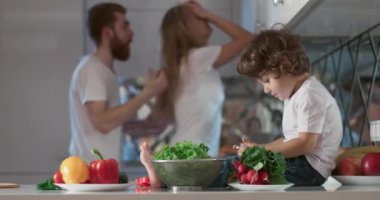 Oğulları yanlarında oturmuş telefonda oyun oynarken mutfakta tartışan genç bir çiftin portresi. Ayrıca ön planda farklı sebzeler ve meyveler vardır. 4K
