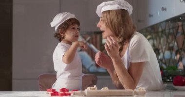 Gülümseyen genç annenin portresi mutfakta oğluyla yemek pişiriyor, kirli elleriyle, pişirmek için para kazanıyor, yaşam tarzı için. Anne ve bebek birbirlerine un sürerler. Beyaz, rahat bir mutfak.