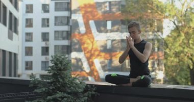 Spor giyimli genç adam lotus pozunda oturuyor. O, balkonda yoga egzersizleri yapıyor. 4K video