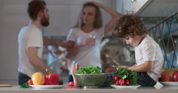 Portrait de jeunes parents se disputant à la cuisine tandis que leur fils est assis à côté d'eux et les regardant. En outre, au premier plan se trouvent différents légumes et fruits comme décoration. Vidéo 4K — Video