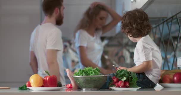 Porträt junger Eltern, die sich in der Küche streiten, während ihr Sohn neben ihnen sitzt und sie ansieht. Im Vordergrund stehen auch verschiedene Gemüse- und Obstsorten als Dekoration. 4K-Video — Stockvideo