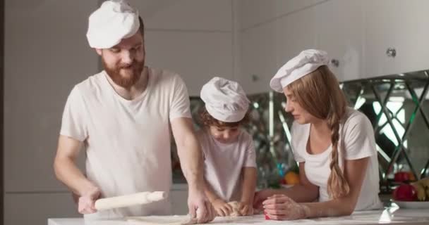 Potret keluarga muda yang tersenyum sedang memasak di dapur. Mereka berlutut adonan dan bersenang-senang. Dapur yang nyaman dan suasana keluarga yang tenang. Video 4K — Stok Video