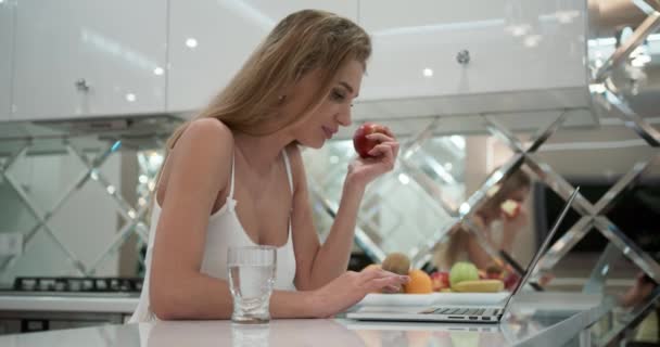 Krásná mladá žena sedí na stole v bílé, útulné kuchyni, jí jablko a hledá něco na svém notebooku. Sexy žena v lákavém pyžamu. Útulný domácí interiér. Video 4K
