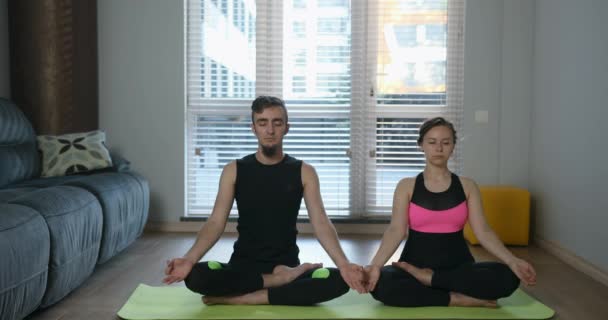 Man och kvinna sitter i lotusställning. Moderna unga par i sportkläder gör yogaövningar på mattan i vardagsrummet hemma. Mysigt rymligt rum och trevlig atmosfär. 4K-video — Stockvideo
