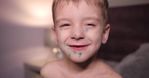 Ritratto di un ragazzo felice malato di varicella. Il bambino guarda dritto nella telecamera e sorride. un paziente con la varicella si siede a casa su un letto. dietro il ragazzo luce fioca dalla lampada del comodino. — Video Stock