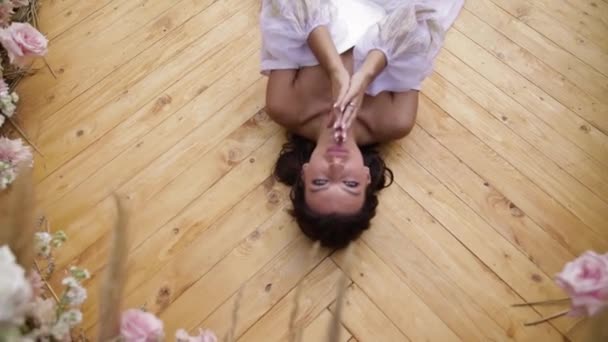 Bride lying on the wooden ground among beatiful flowers, in a wedding dress that touches a curl of her hair, slow motion — Stock Video