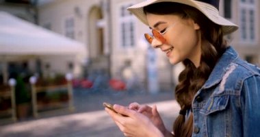 Kot ceketli ve pembe yuvarlak gözlüklü genç kadın şehir merkezinde akıllı telefon kullanıyor ve gülümsüyor..