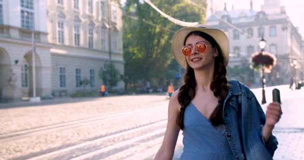 Young woman in a blue dress, a straw hat, pink round glasses, a denim jacket, walks the city center. A tram rides behind and people walk. Woman is happy and smiling