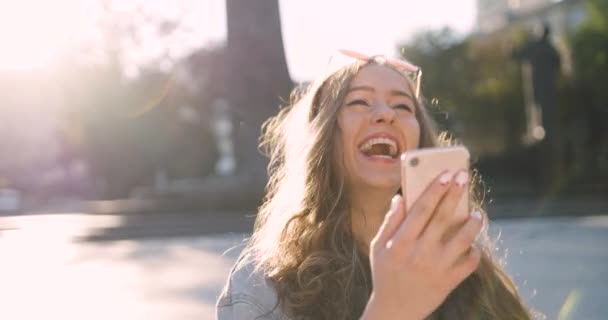 Menina atraente com cabelo loiro longo olha para a tela do smartphone, fala em bate-papo por vídeo e ri sinceramente. — Vídeo de Stock