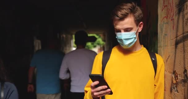 Um jovem fica na rua com uma máscara protetora com um telefone na mão encostado à parede — Vídeo de Stock