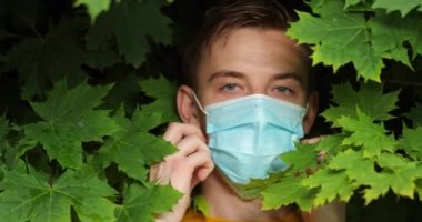 Yüzünde çiller olan çekici bir adam yeşil yaprakların arasında duruyor kameraya bakıyor ve koruyucu tıbbi maskeyi çıkarıyor.