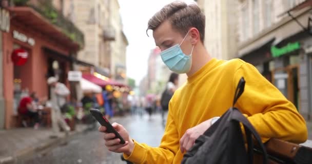 Mladý muž v pozadí budovy pod širým nebem s maskou, která chrání před rozšířením koronavirové choroby. Detailní záběr muže s chirurgickou maskou na tváři — Stock video
