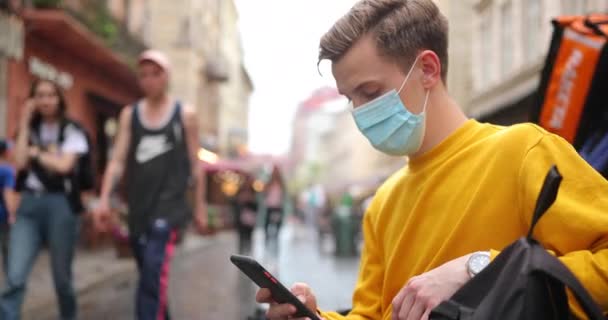 Běloch městský kluk student s červenými vlasy psaní na smartphone zprávy při chůzi po ulici s ochrannou maskou proti koronaviru. Pandemie — Stock video