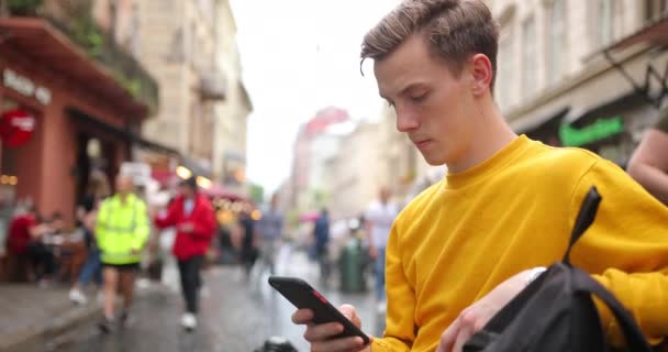 Cara loiro atraente fica na rua em uma cidade onde muitas pessoas e usa um smartphone — Vídeo de Stock