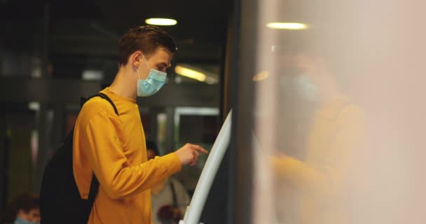 A guy in a yellow sweater and a black backpack is standing in a medical mask near the bank terminal — Stock Video