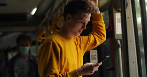 Männlich in öffentlichen Verkehrsmitteln ohne Schutzmaske benutzte Smartphone. Junger attraktiver Mann im gelben Pullover steht im Bus — Stockvideo