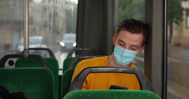 Молодий хлопець їде в автобусі з медичною маскою на обличчі. Захист здоров'я під час епідемії. Молодий чоловік їздить на порожньому автобусі в медичній масці і користується телефоном. Хлопець у масці сидить у автобусі — стокове відео