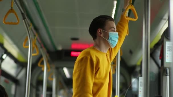 Здоров'я та безпека під час глобальної епідемії коронавірусу. Безпека на громадському транспорті. Хлопець їде на автобусі в захисній медичній масці, тримаючись за поручні і дивлячись у вікно — стокове відео