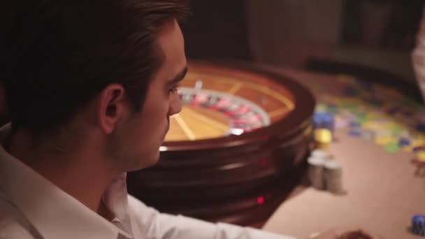 Nervöser brünetter Mann im weißen Hemd trinkt Whiskey und sitzt allein hinter dem Roulettetisch im Casino, während der Dealer alle Chips vom Tisch nimmt. Wütender Mann nach Niederlage im Casino — Stockvideo