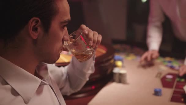 De cerca. Nervioso morena hombre en camisa blanca beber whisky y sentado solo detrás de la mesa de ruleta en el Casino mientras distribuidor se lleva todas las fichas de la mesa — Vídeo de stock