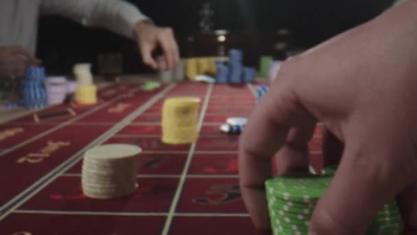 De cerca. Mesa de ruleta con manos humanas poniendo fichas en el casino. Personas irreconocibles se sientan detrás de la mesa de juego, bebiendo whisky y fumando cigarros mientras hacen apuestas — Vídeo de stock