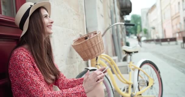 Mujer joven en un sombrero con una bicicleta sentada en la calle cerca de la puerta roja. Gente activa. Al aire libre. — Vídeos de Stock