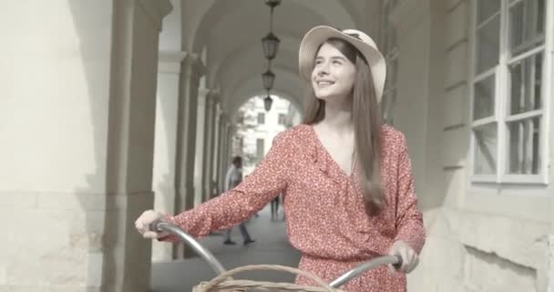 Chica con una bicicleta pasea por la ciudad y camina cerca del estacionamiento de bicicletas. detrás del arco. clima cálido y soleado — Vídeo de stock
