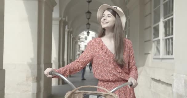 Милая девушка в красном платье и соломенной шляпе идет вдоль велосипеда по улице города — стоковое видео