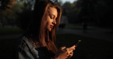 Uzun sarı saçlı çekici kız parktaki akıllı telefon ekranına bakıyor..
