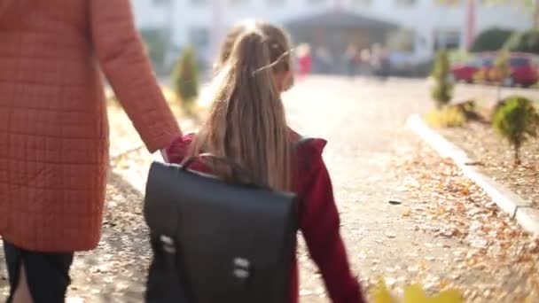 Matka i dziewczynka w czerwonej sukience z plecakiem na ramionach i bukietem żółtych jesiennych liści idą do szkoły. Słoneczna jesień. Wideo 4K. — Wideo stockowe