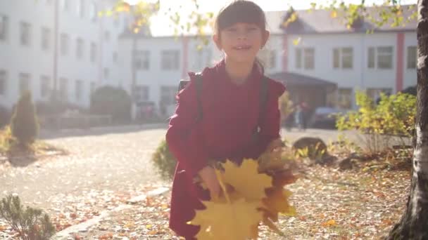 Holčička v červených šatech s batohem na ramenou zvrací na dvorku školy kytici žlutých spadlých listů. Slunečný podzim. Video 4K. — Stock video