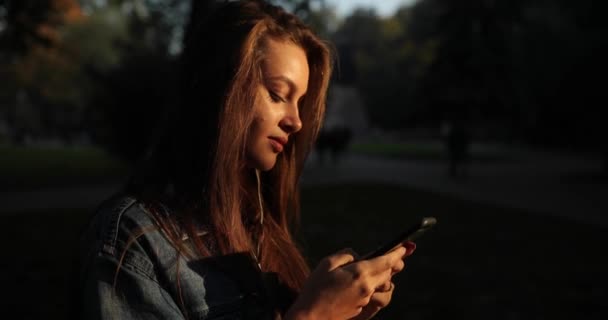 Menina atraente com cabelo loiro longo olha para a tela do smartphone no parque. — Vídeo de Stock