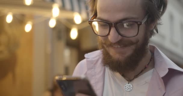 4k. Hipster turista masculino na cidade. Homem barbudo de óculos caminha ao longo da velha rua europeia e lê algo em seu smartphone. Emoções positivas — Vídeo de Stock