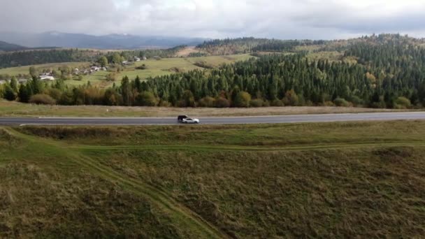 Mobil menyalip di jalan dengan latar belakang pegunungan, hutan dan desa. Tembakan quadcopter. SUV vs sedan. — Stok Video