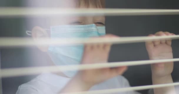 Un niño con una máscara médica en la cara mira por la ventana y estornuda. Vista exterior del niño molesto con máscara mirando a la ventana durante el bloqueo de cuarentena para Coronavirus Covdi19. Autoaislamiento. — Vídeos de Stock
