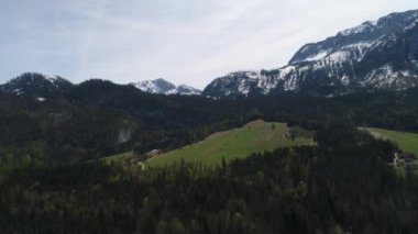 Avustralya köyünün havadan görünüşü ve baharda Alpler 'in karlı zirveleri. Yeşil orman, tarla, beyaz gökyüzü ve güneşli gün. Avrupa. High Mountain Range 'den panoramik manzara. 4K video.