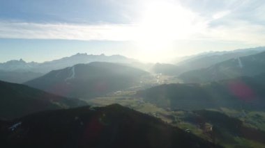 Avustralya köyünün panoramik manzarası ve ilkbaharda güneşin batışında sisli Alpler 'in karlı tepeleri. Yeşil orman, tarla, beyaz gökyüzü ve güneşli gün. Avrupa. High Mountain Range 'den insansız hava aracı görüntüsü. 4K video.