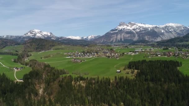 Vue par drone du village autrichien et des sommets enneigés des Alpes au printemps. Forêt verte, champ, ciel blanc et journée ensoleillée. L'Europe. Vue panoramique depuis la haute montagne. Vidéo 4K. — Video