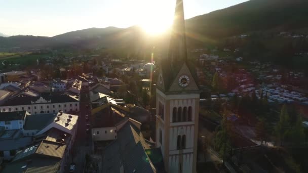 Sunset view of beautiful landscape of small spring town among Austrian alpine mountains. Drone video of church and small houses on the background of mountains easily covered with snow. 4K video. — Stock Video