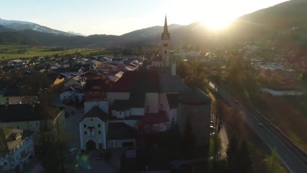 Sunset view of beautiful landscape of small spring town among Austrian alpine mountains. Drone video of church and small houses on the background of mountains easily covered with snow. 4K video. — Stock Video