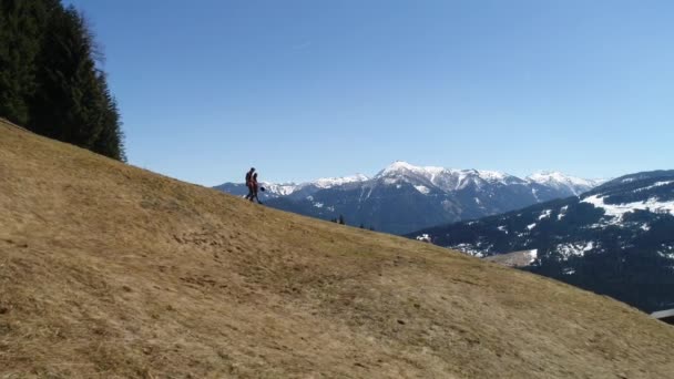 Video vesnického muže se sekerou ukazuje cestu k atraktivní brunetce a ona sestupuje z hory. Rakouská vesnice a zasněžené vrcholky Alp na jaře. Zelený les, pole, bílá obloha — Stock video